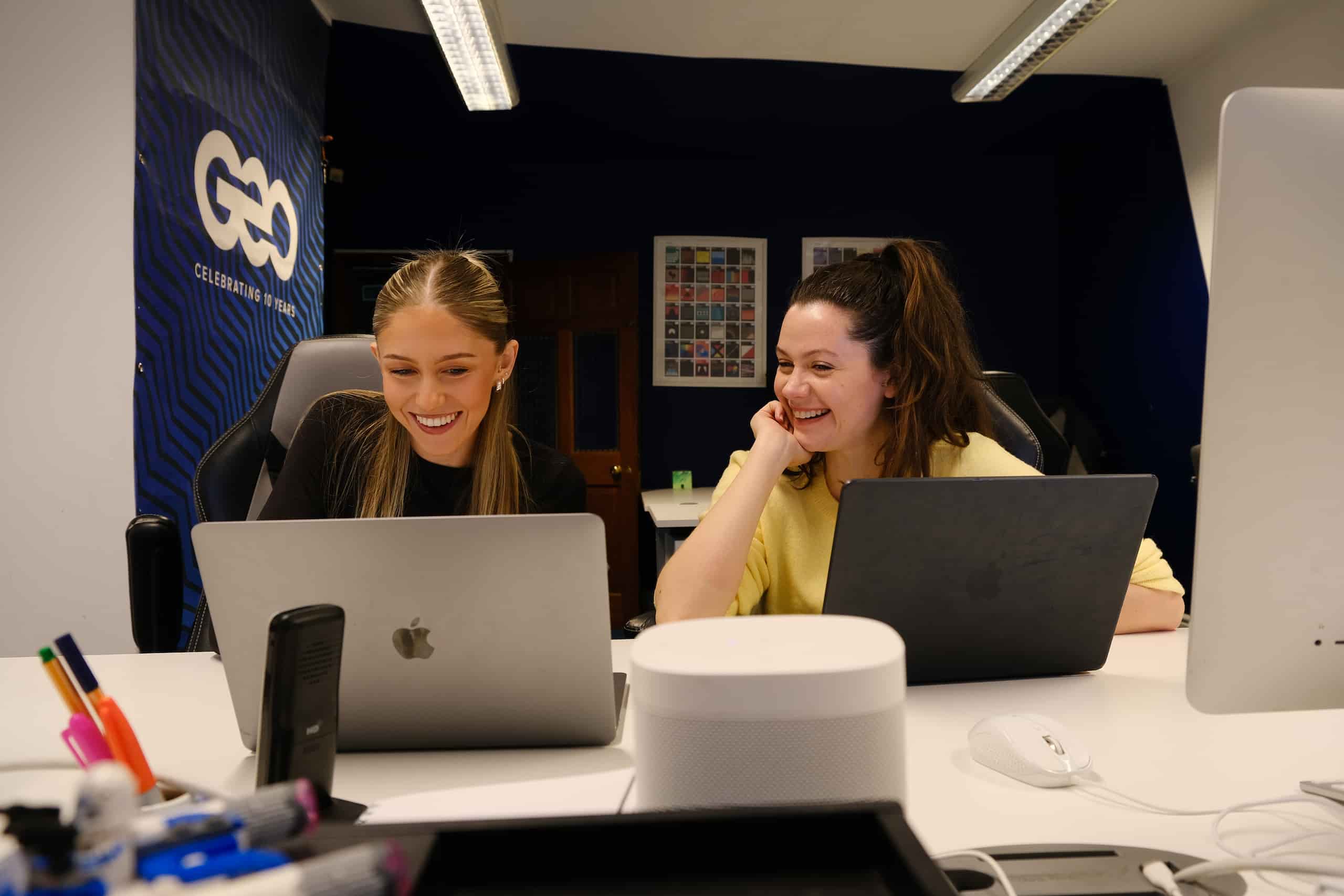 Ellie and Caroline Working Together Ellie and Caroline smiling while working together on laptops at a desk in a modern creative office.
