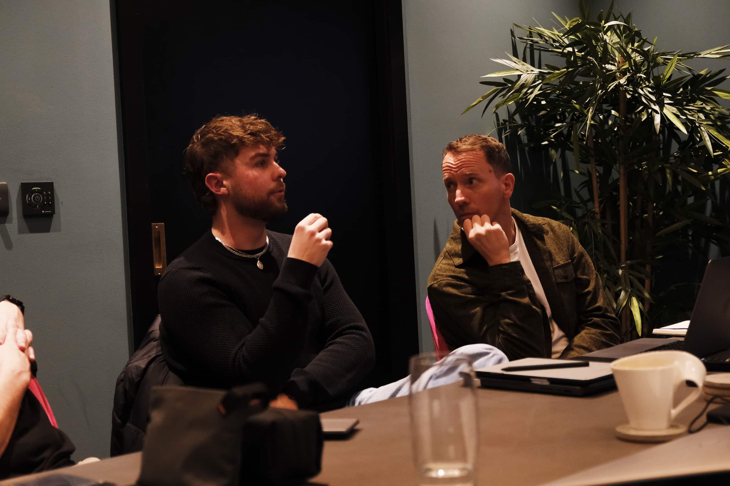 Kian and Andrew engaged in discussion during a meeting, seated at a table with laptops in a modern office environment.