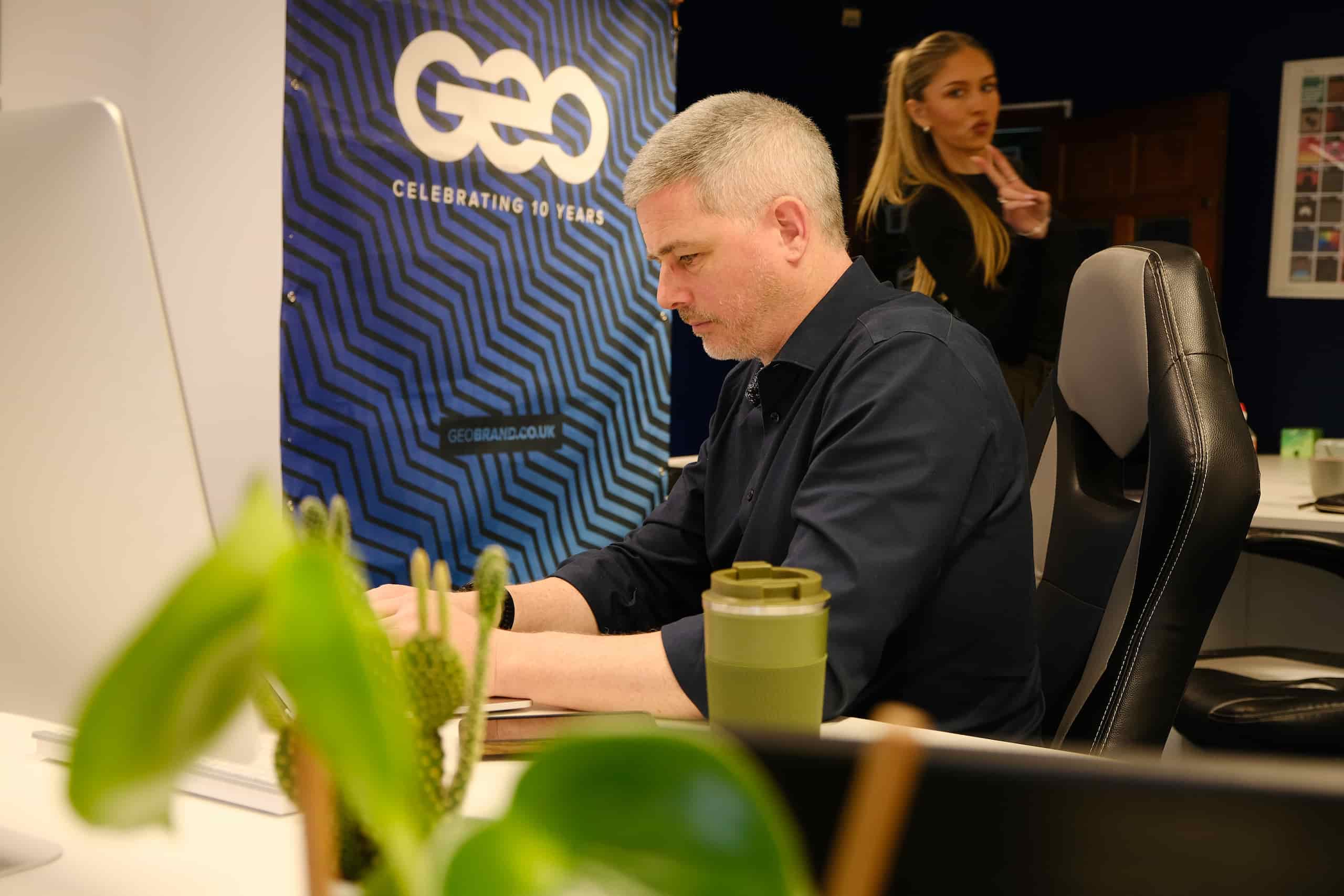 Lee working at a desktop computer while Ellie stands in the background in a modern office workspace.